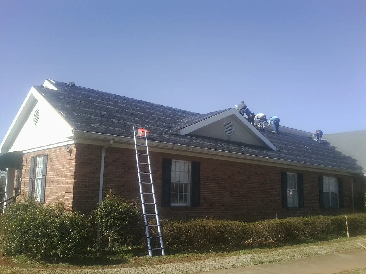 Crew tearing shingles off a brick ranch roof with a ladder set — Metal Roof Repair work by our crew in Austinburg
