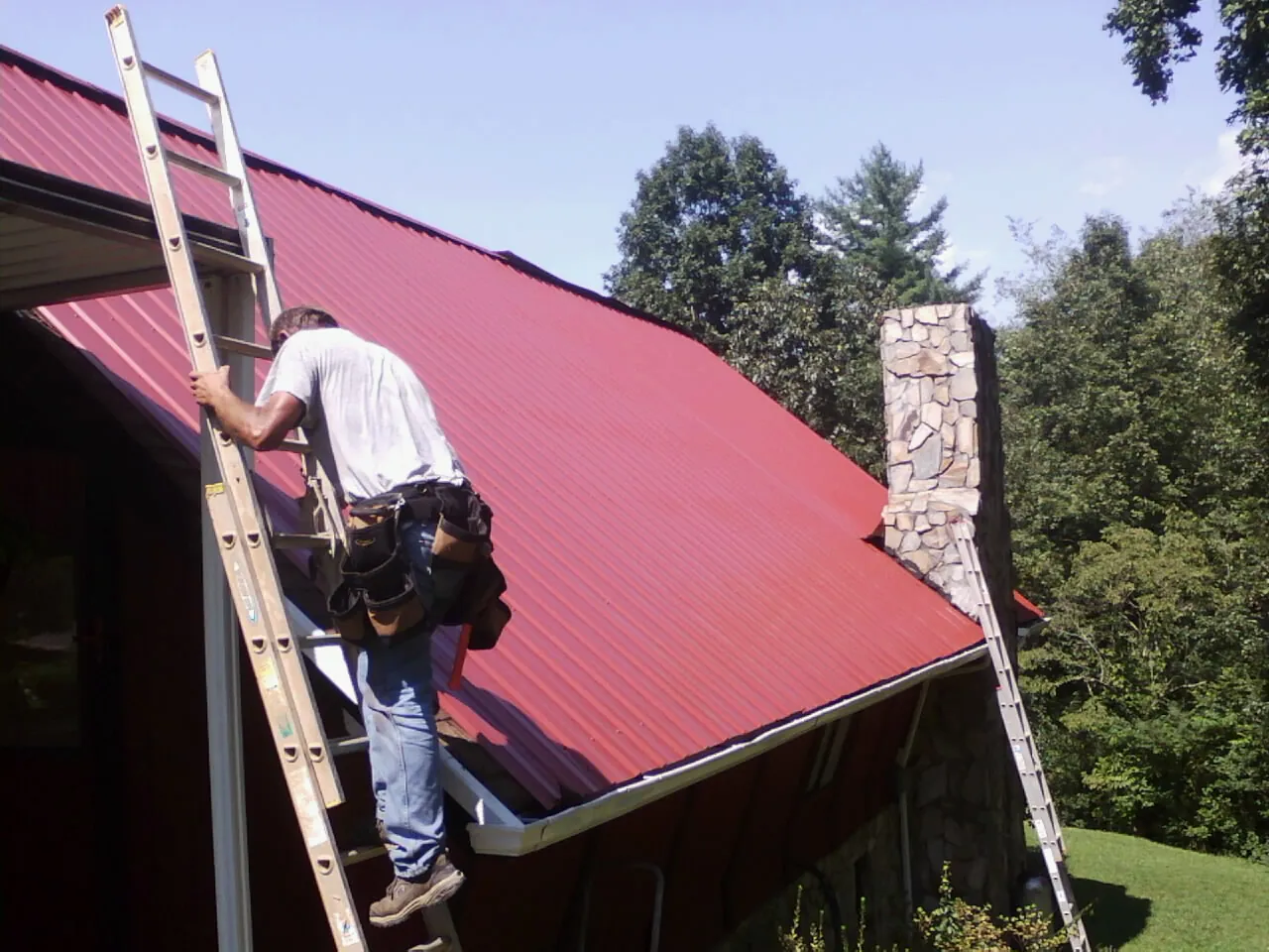 Roofer on a ladder beside a red metal roof and stone chimney — Soffit & Fascia Repair by Roofing Brothers in Austinburg